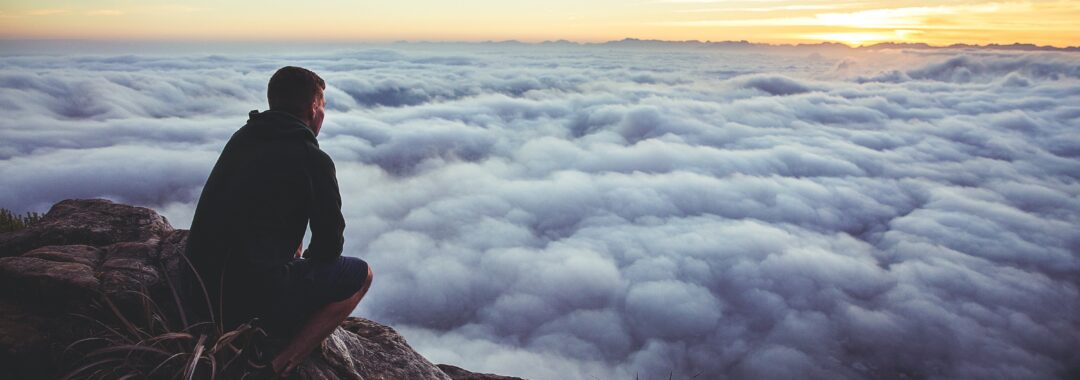 man sitting on gray rock while staring at white clouds