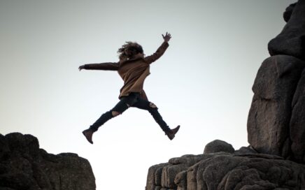 person jumping on big rock under gray and white sky during daytime