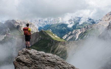 man with red backpack standing on cliff facing mountains under white sky during daytime