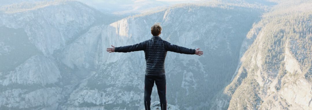 man opening his arms wide open on snow covered cliff with view of mountains during daytime